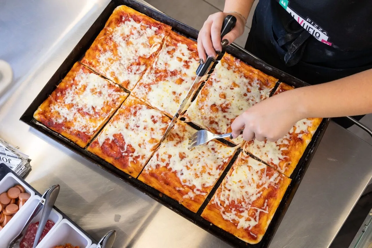 Dos manos cortan en porciones rectangulares la pizza recién horneada, todavía en la bandeja.