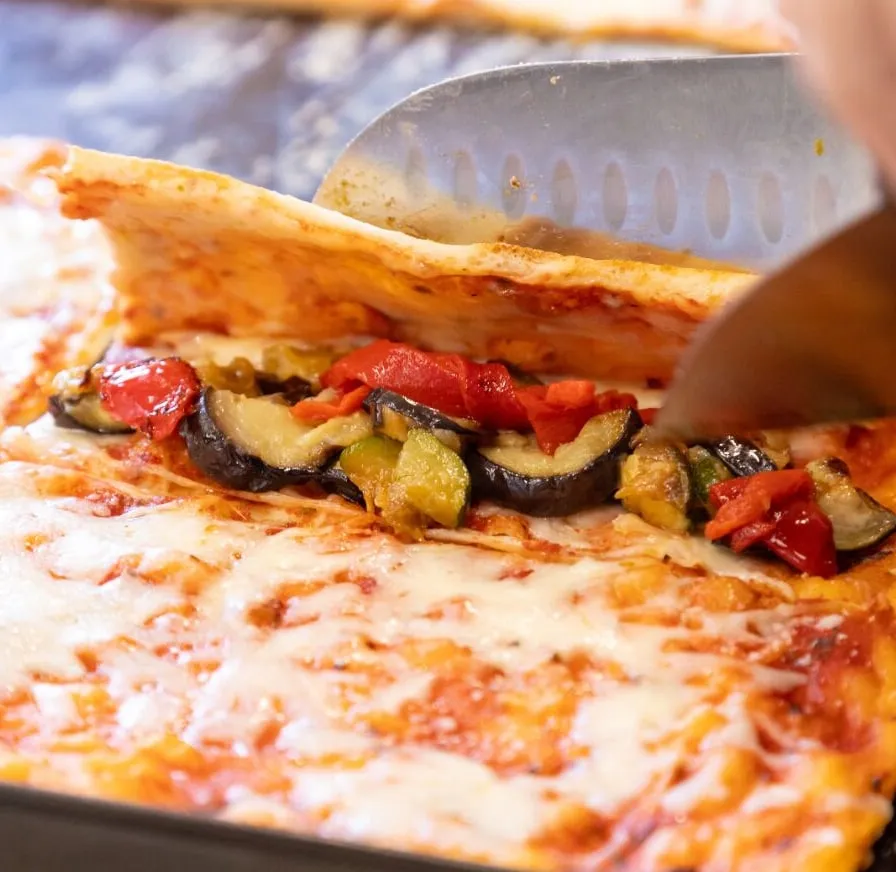 Un cuchillo dobla una porción de pizza sobre un relleno de berenjena, calabacín y pimiento rojo.