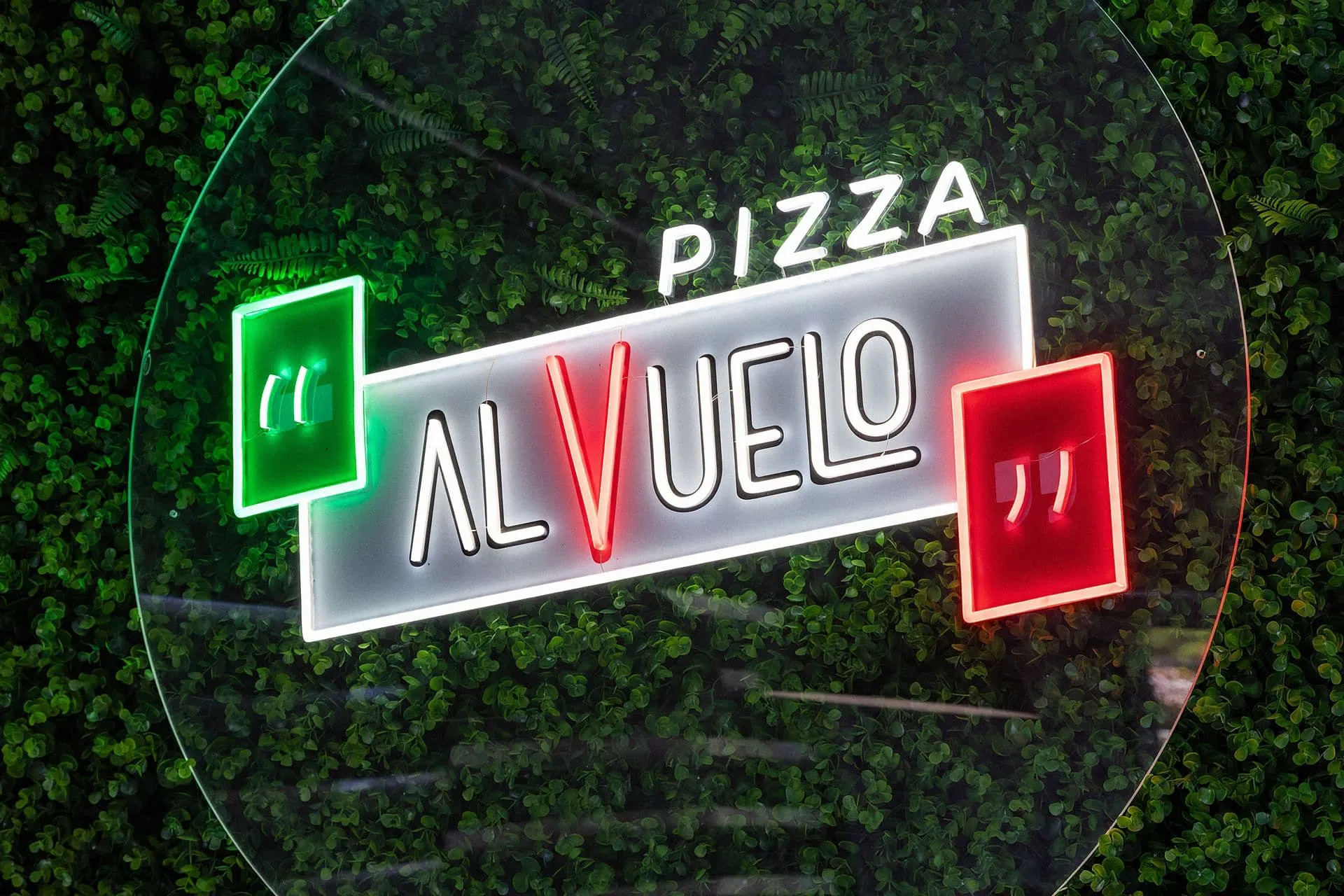 Letrero de neón de Pizza al Vuelo, con las comillas verde y roja, sobre una pared de plantas.