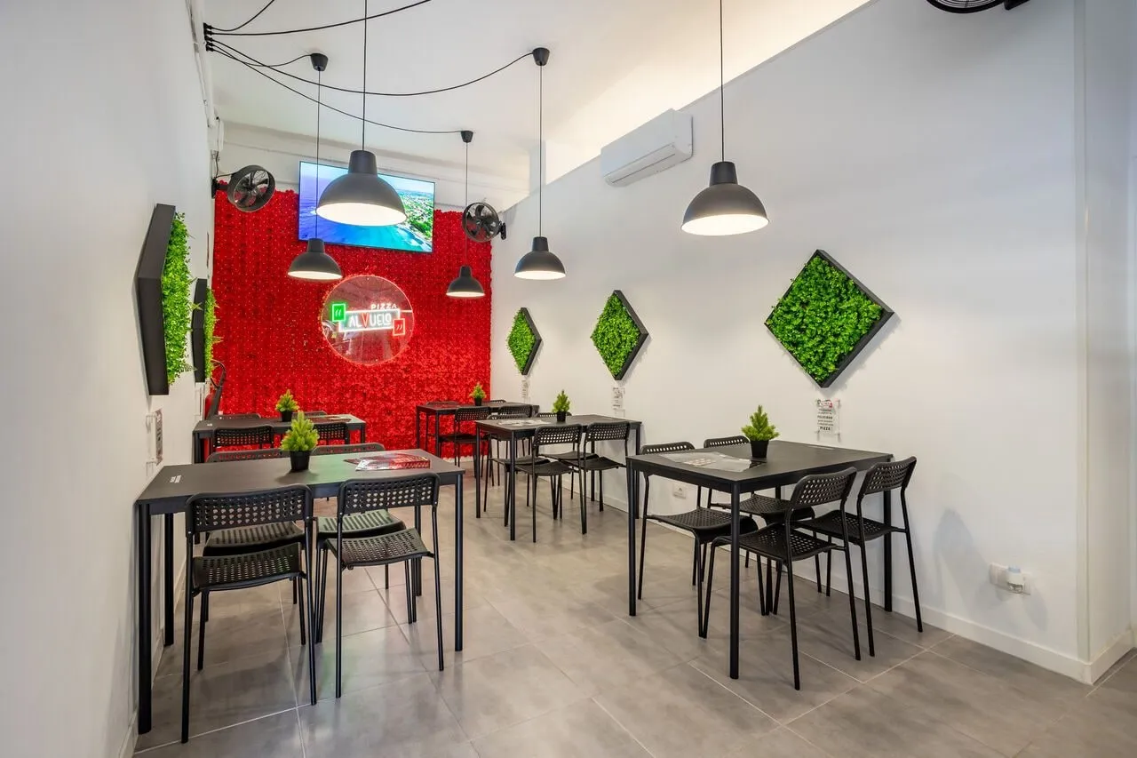 Sala de Pizza al Vuelo: pared roja con el logo luminoso, mesas y sillas negras, lámparas colgantes y rombos de musgo.